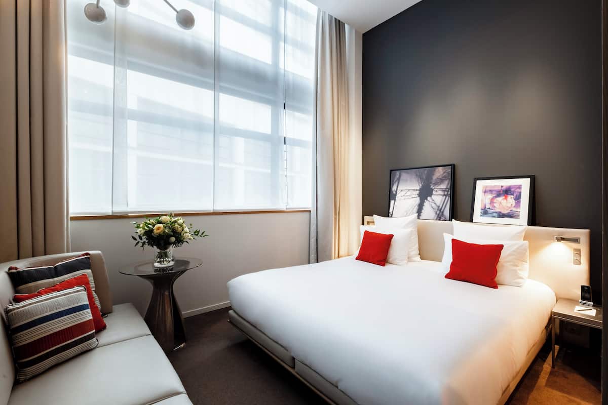 A hotel room with a large bed, a sofa, a small table with a vase of flowers, and artwork on the wall.