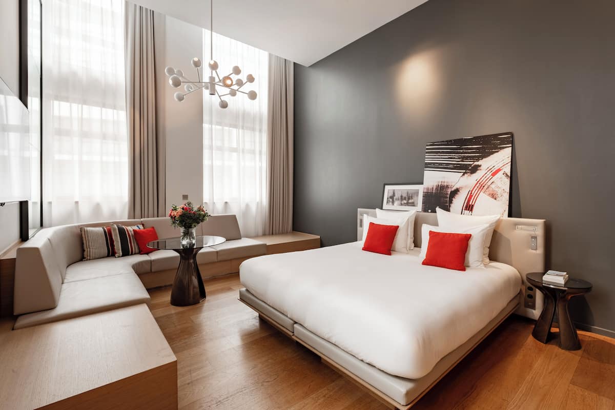 A modern hotel room with a large bed, a sofa, a small table, and a large window with curtains.