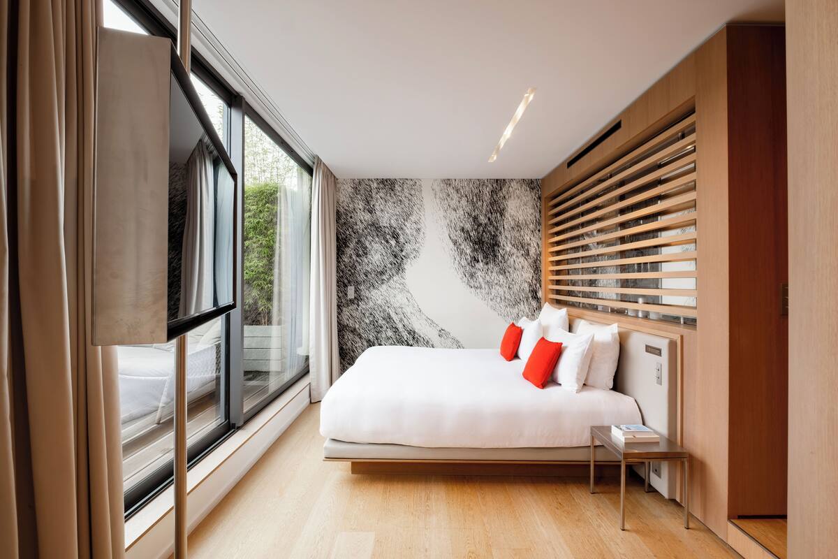 A modern bedroom with a large bed, a bedside table, and a view through a window.