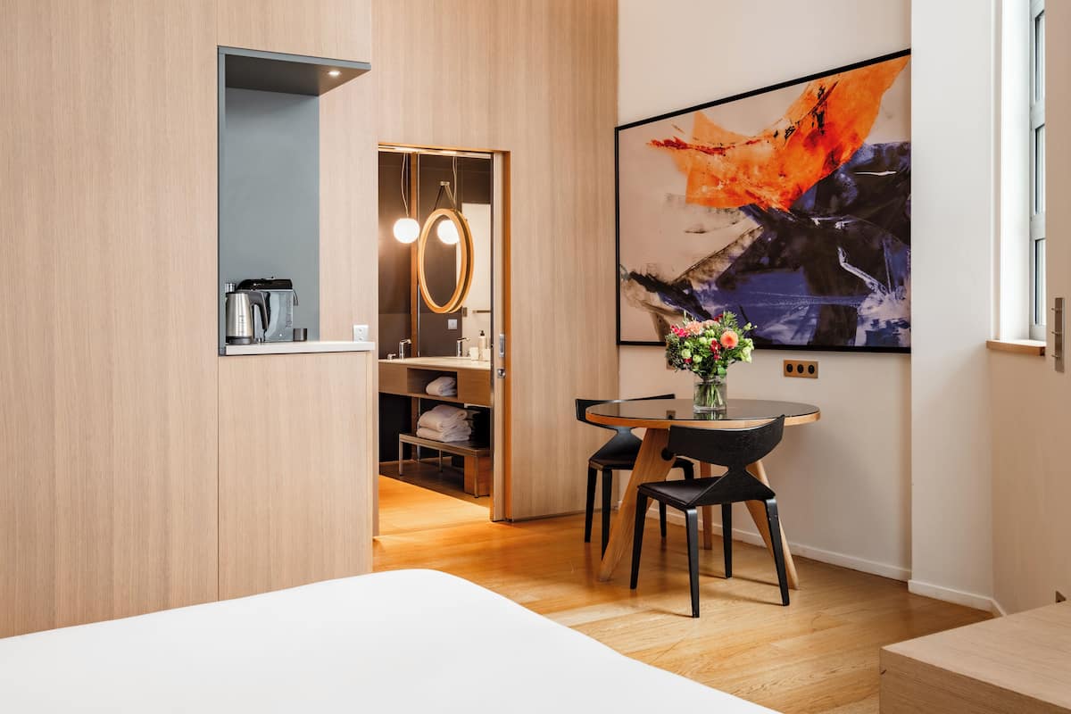 A modern hotel room with a wooden floor, a bed, a dining table with chairs, a coffee machine, a mirror, and a large abstract painting.