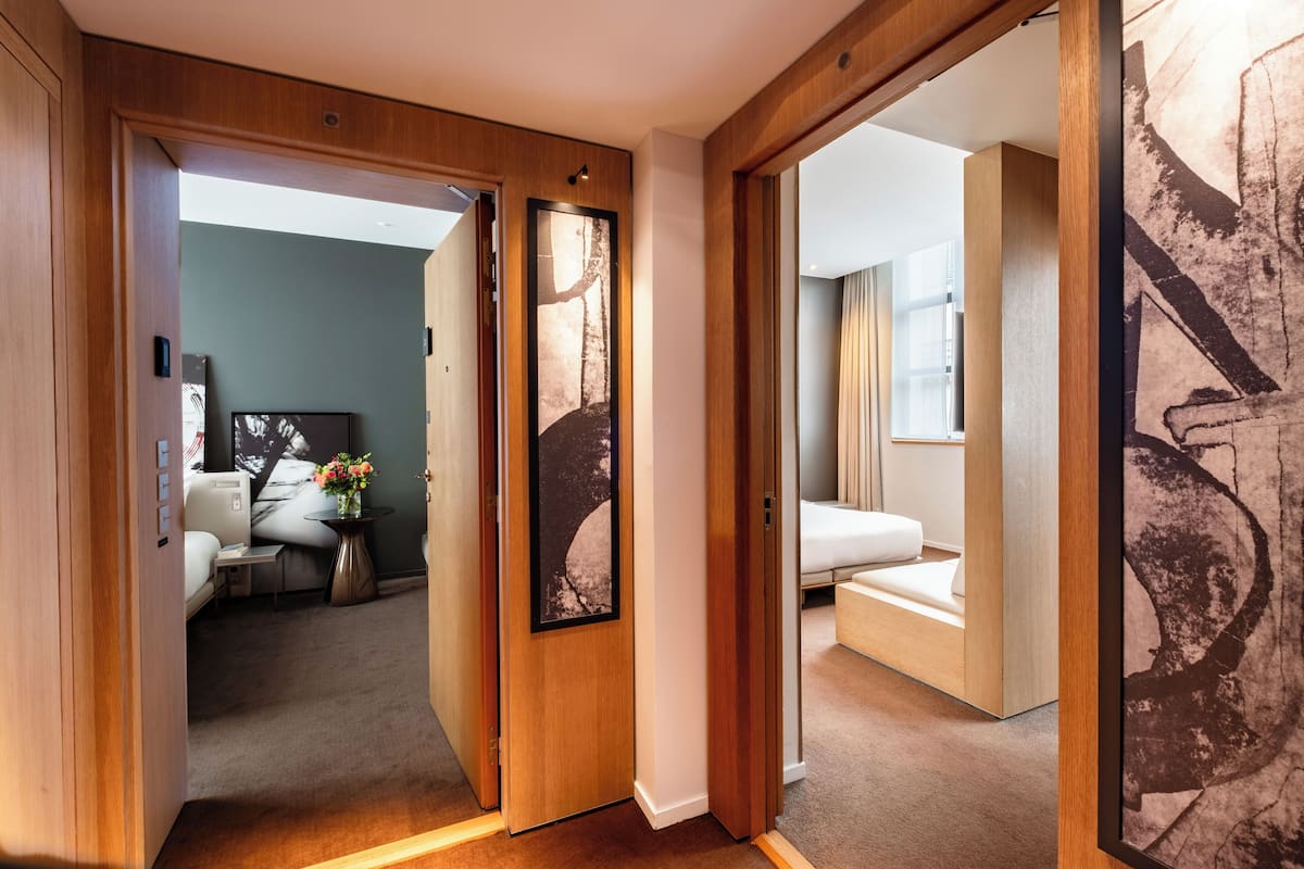 A hotel room with a wooden door, a bed, a small table with a vase, and a large abstract painting on the wall.