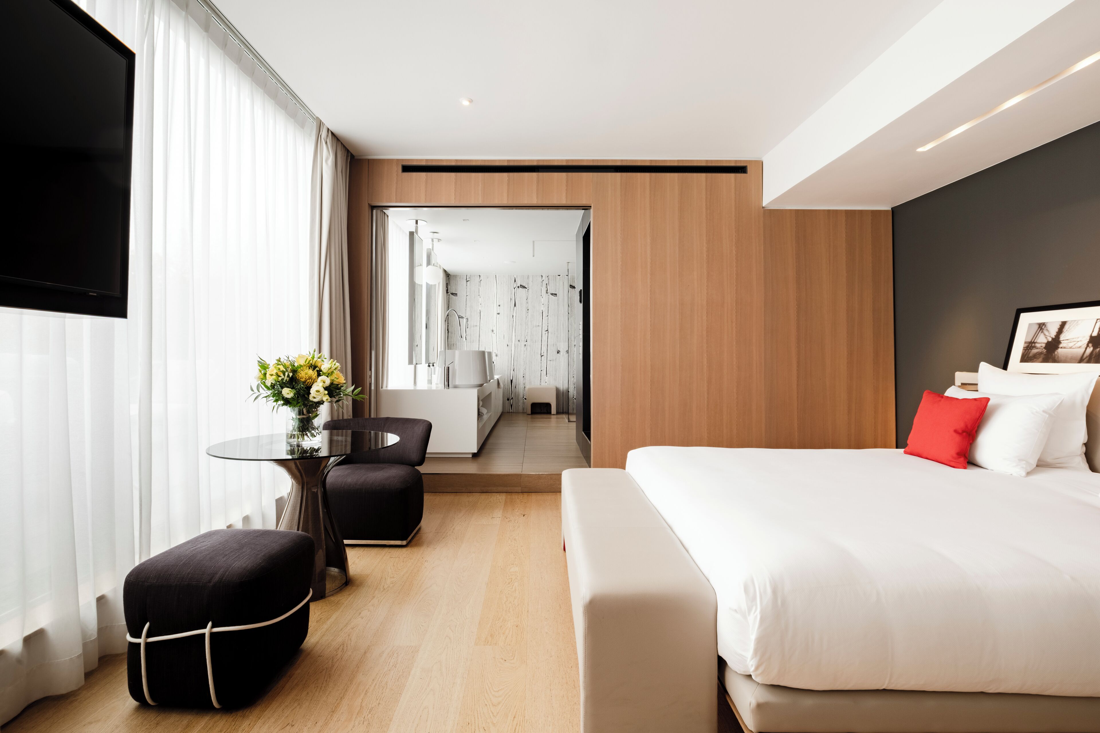 Une chambre d’hôtel moderne avec un grand lit, une petite table ornée d’un vase de fleurs, un coin salon avec un fauteuil et un téléviseur fixé au mur.
