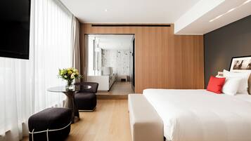 Une chambre d’hôtel moderne avec un grand lit, une petite table ornée d’un vase de fleurs, un coin salon avec un fauteuil et un téléviseur fixé au mur.