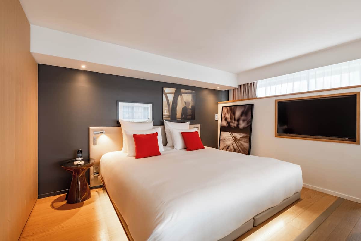 A modern hotel room with a large bed, a bedside table, a nightstand, and a large window.