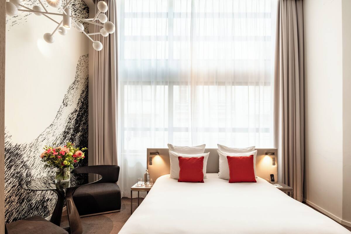 A hotel room with a large bed, two red pillows, a bedside table with a lamp, a chair, and a wall with a modern abstract design.
