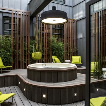 A modern outdoor hot tub area with wooden decking, green chairs, and a large overhead light.