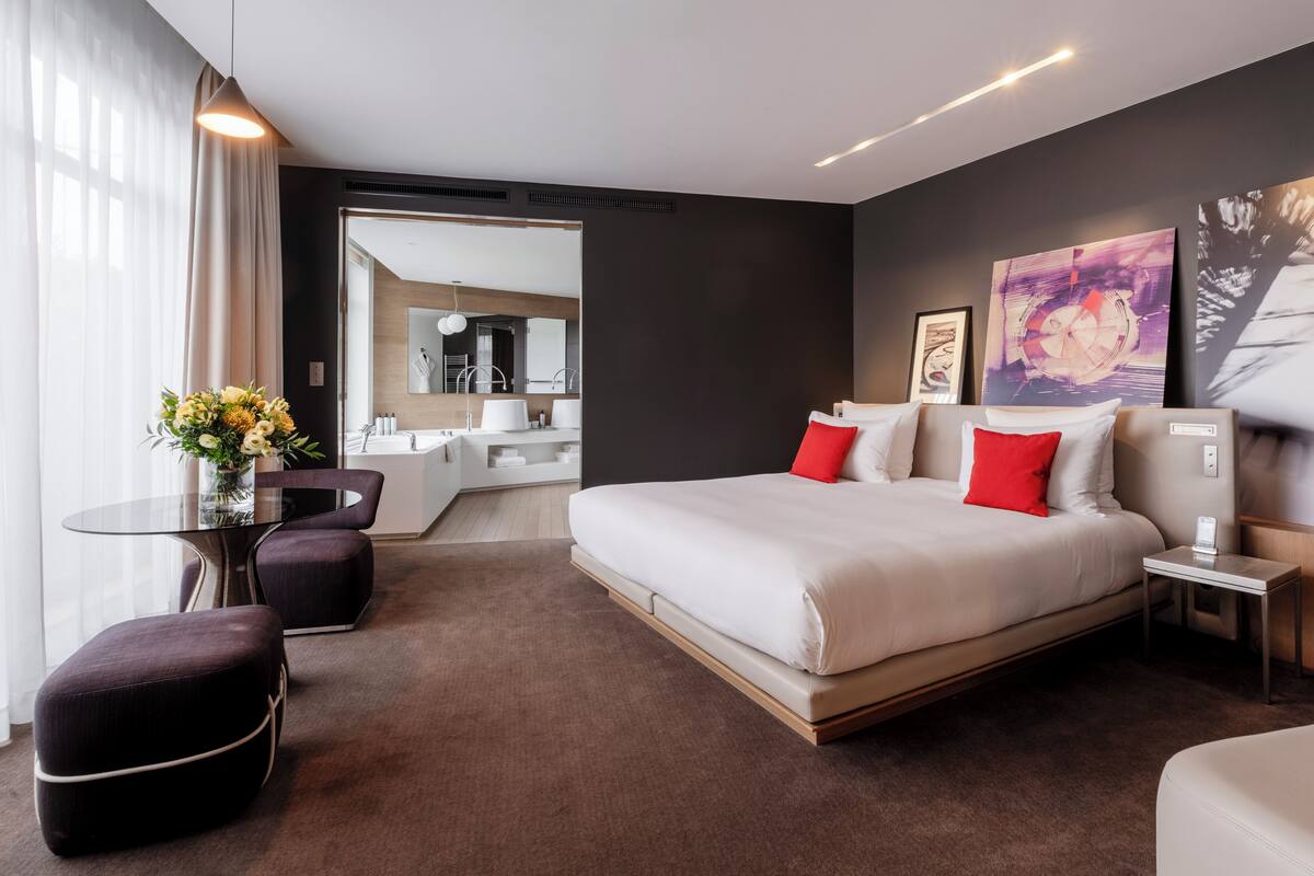 A modern hotel room with a large bed, a sitting area with a table and chairs, a small kitchen area, and artwork on the walls.