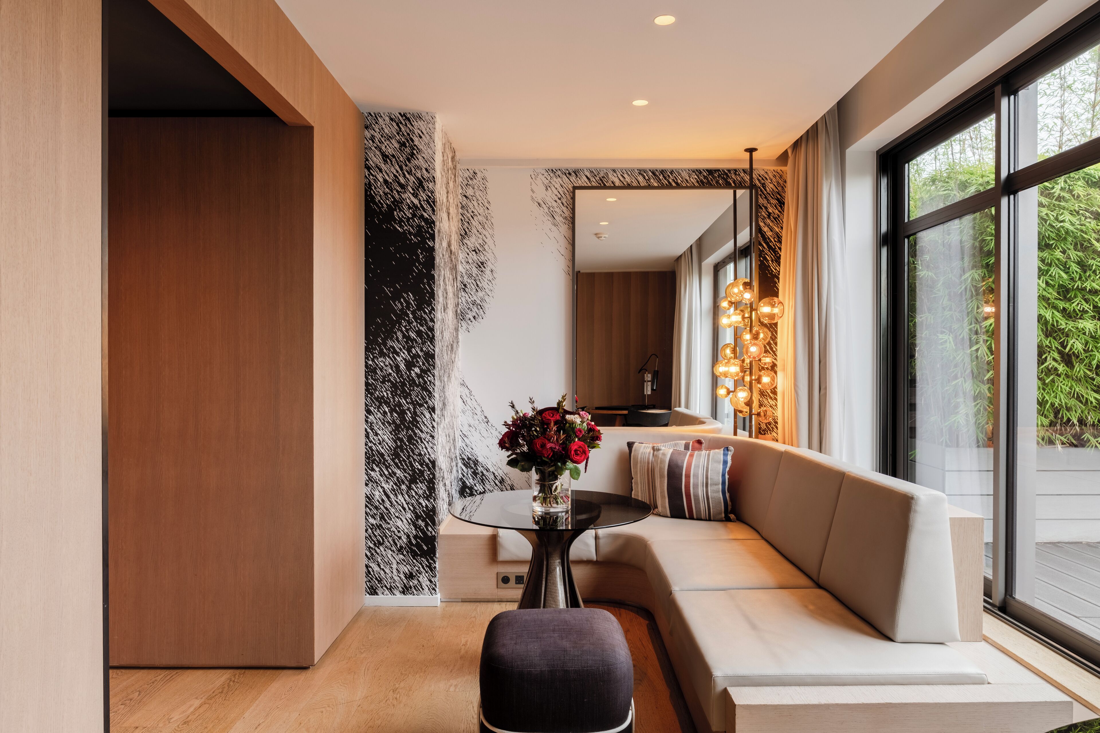 Ein modernes Interieur mit Holzboden, einer strukturierten Wand in Schwarz-Weiß, einem runden Tisch mit Blumenvase, einer Couch mit Kissen und einem großen Fenster mit Blick auf Grünflächen.