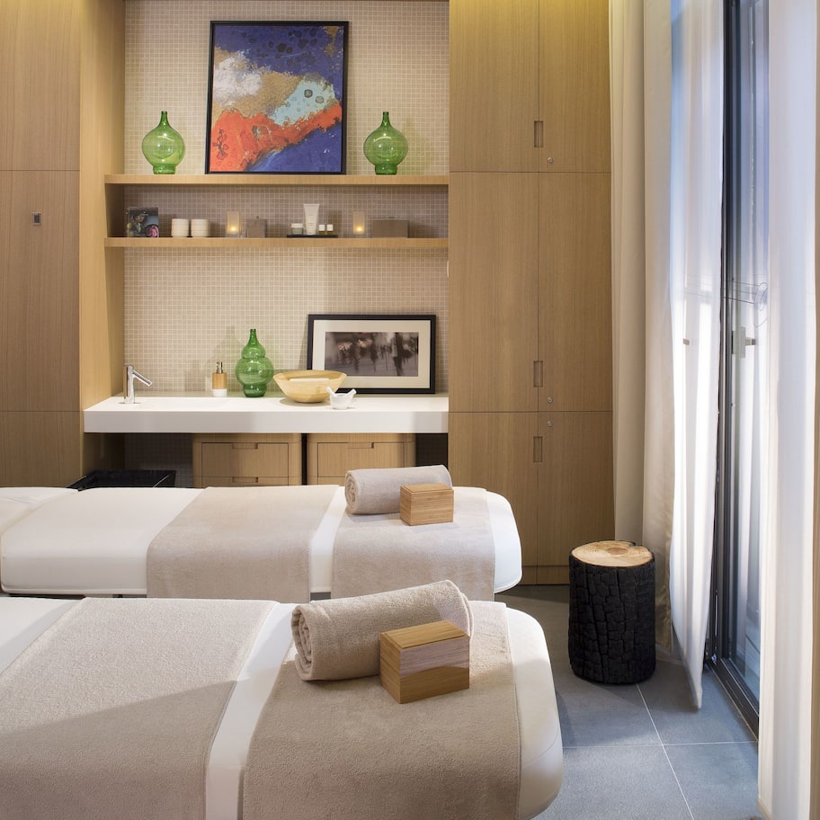 A modern spa room with two massage beds, a small sink, and a window with sheer curtains.