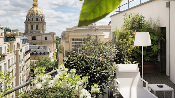 A rooftop garden with a white lounge chair, a table, and a lamp. In the background, there is a large building with a dome and a golden spire. - Le Cinq Codet (Paris)