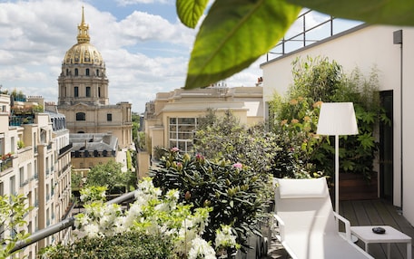 A rooftop garden with a white lounge chair, a table, and a lamp. In the background, there is a large building with a dome and a golden spire.