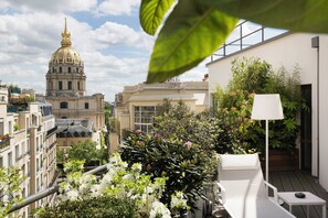 A rooftop garden with a white lounge chair, a table, and a lamp. In the background, there is a large building with a dome and a golden spire.