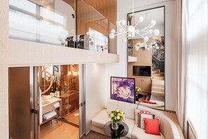 A modern hotel room with a glass-enclosed bathroom, a sofa with a vase of flowers, and a large mirror reflecting the room.