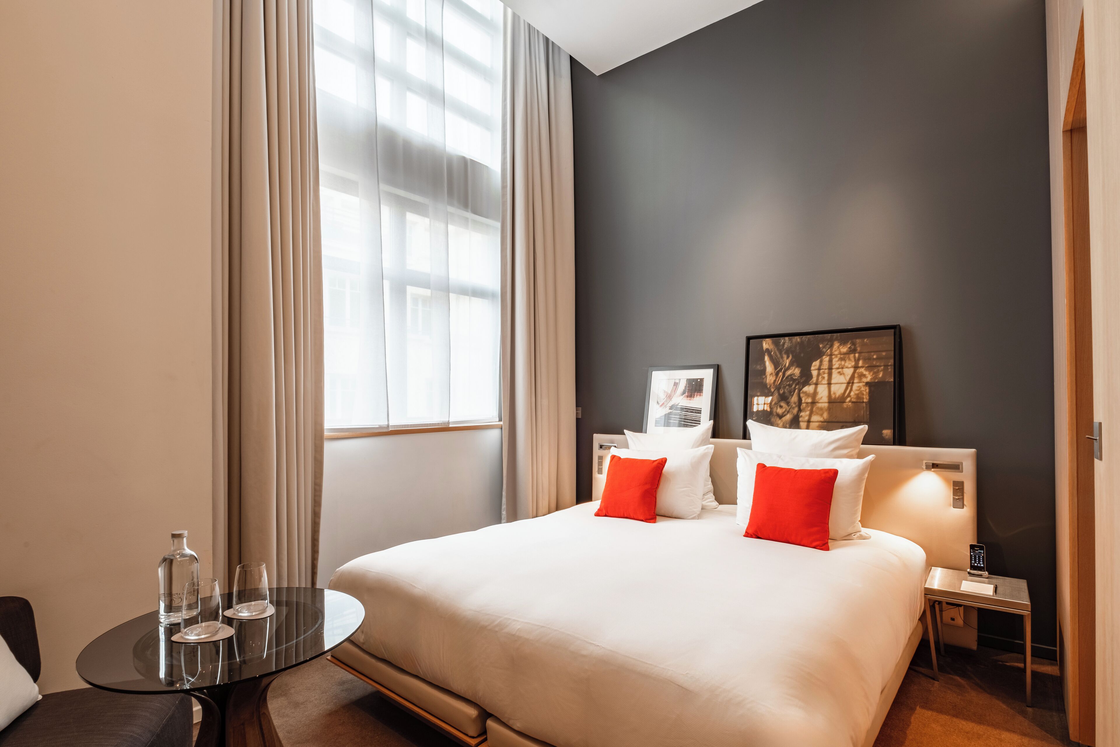 Habitación de hotel con una cama grande, una mesita de noche con lámpara, una pequeña mesa redonda con vasos y una ventana con cortinas.