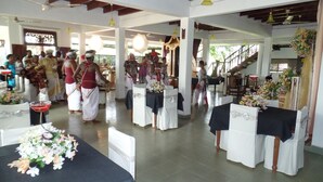 Een ceremonieel evenement met deelnemers in traditionele kledij, tafels met bloemrijke decoraties en een trap die naar een hoger gelegen niveau leidt.