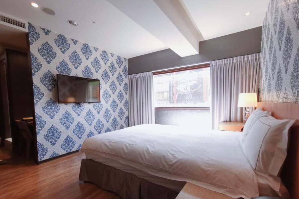 A hotel room with a large bed, a flat-screen TV mounted on the wall, a window with curtains, and a patterned wallpaper.