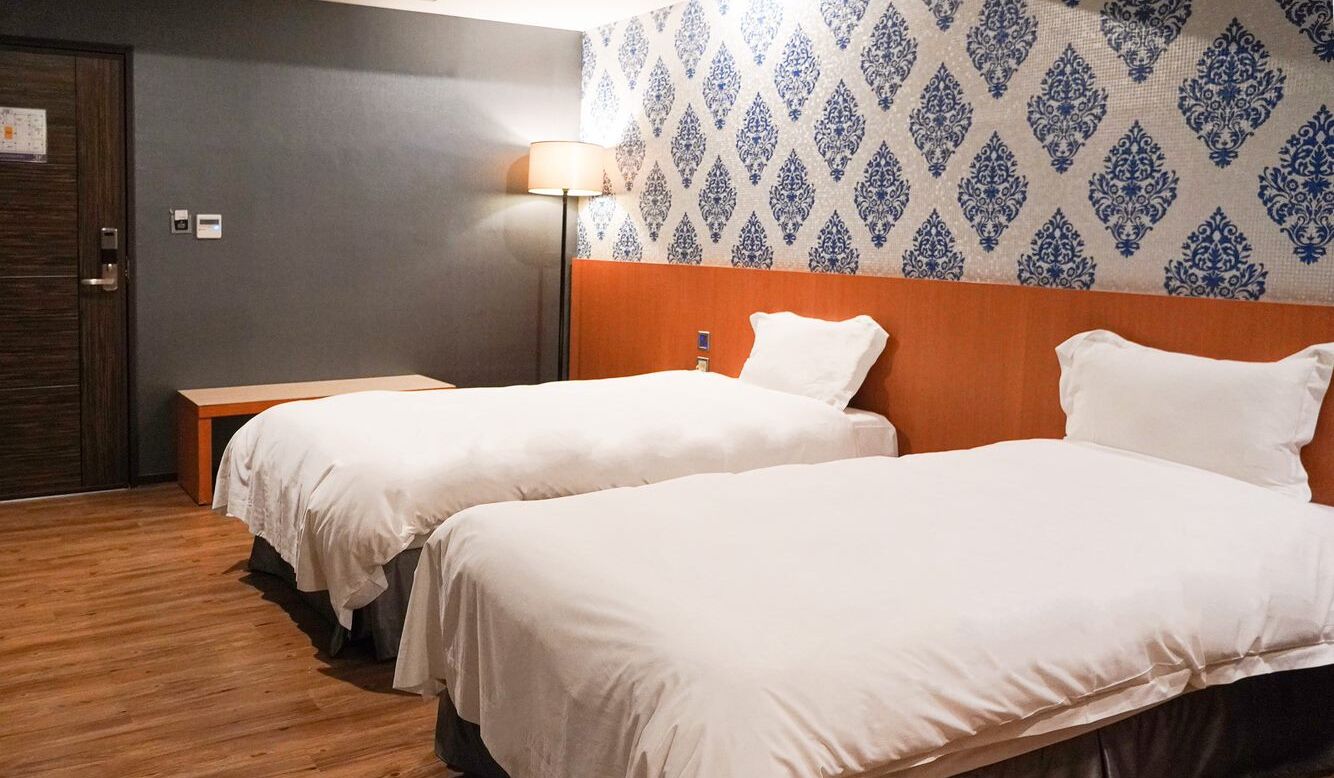 Habitación de hotel con dos camas individuales, suelo de madera, pared con diseño y una mesita de noche con lámpara.