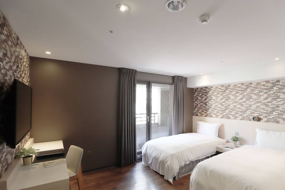 A hotel room with two beds, a desk, a chair, and a large window with curtains.