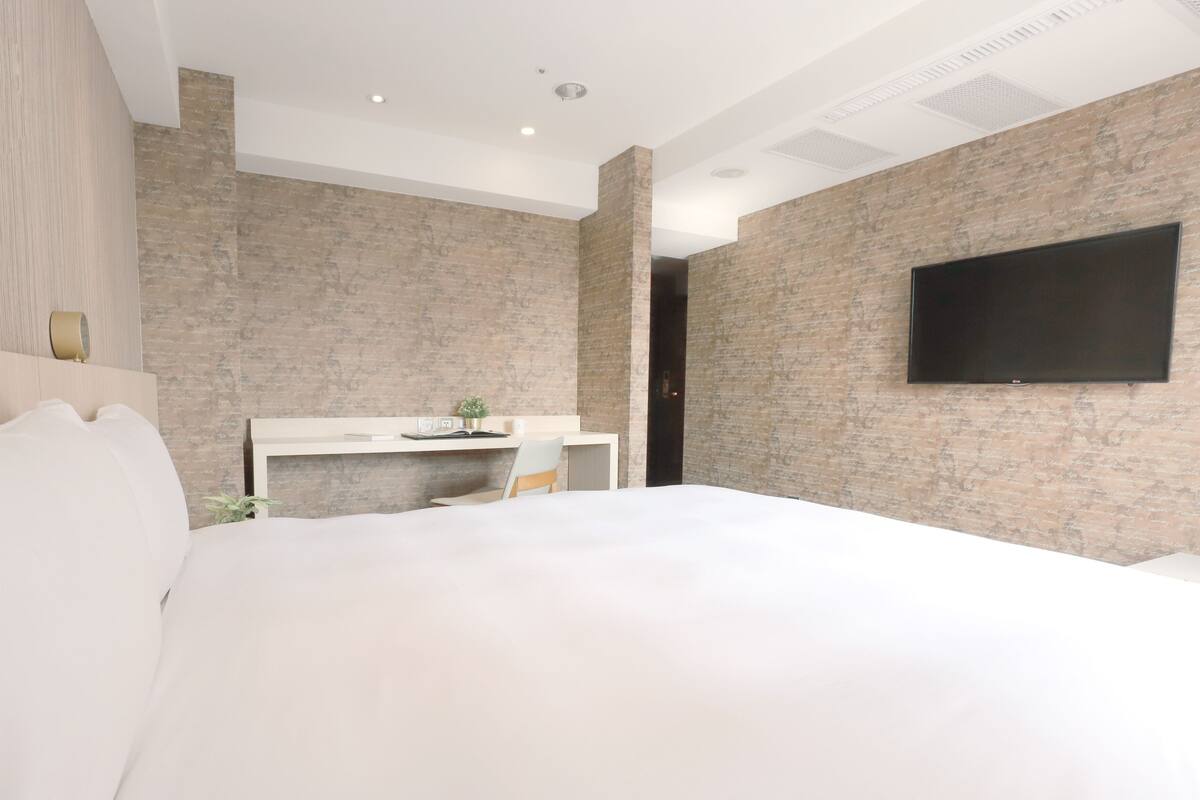 A modern hotel room with a large bed, a desk with a chair, a flat-screen TV mounted on the wall, and a brick-patterned wall.