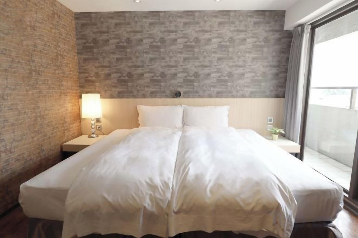 A hotel room with a large bed, a nightstand with a lamp, a view of the outdoors through a window, and a brick wall.