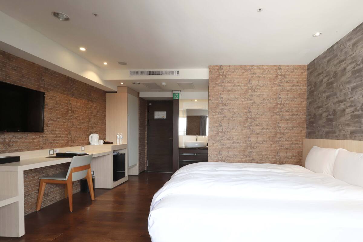 A hotel room with a bed, a desk with a chair, a television, and a bathroom with a sink and mirror.