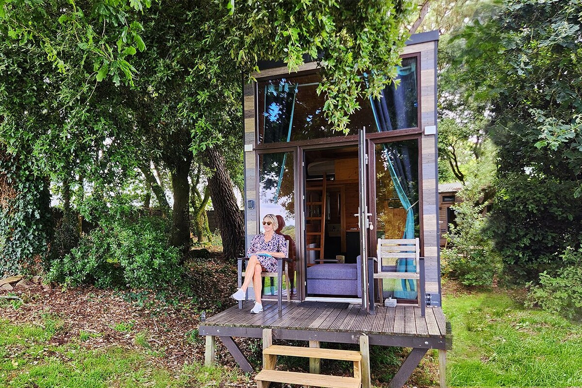Eine Holzterrasse mit einer Bank, eine kleine Holzhütte mit Glastür und eine Person, die auf der Bank sitzt.