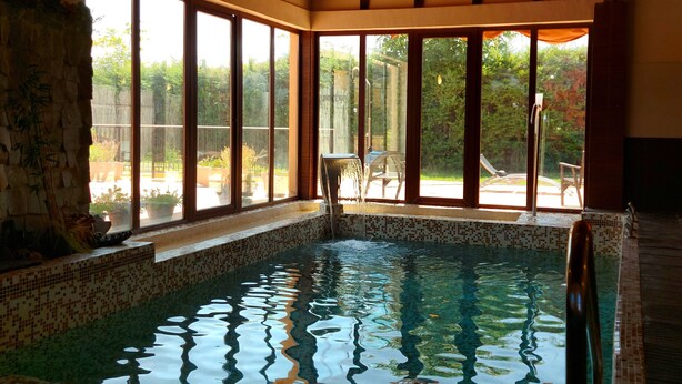 Una piscina interior con paredes de vidrio transparente, vigas de techo de madera y suelo de baldosas.
