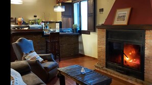 Una sala de estar acogedora con chimenea, comedor con cocina al fondo y un amplio ventanal que deja entrar luz natural.