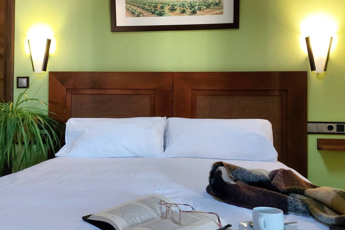 Una cama bien hecha con sábanas blancas, un libro, una taza en un platillo, un cuadro de paisaje enmarcado sobre la cama y dos lámparas de pared a cada lado.