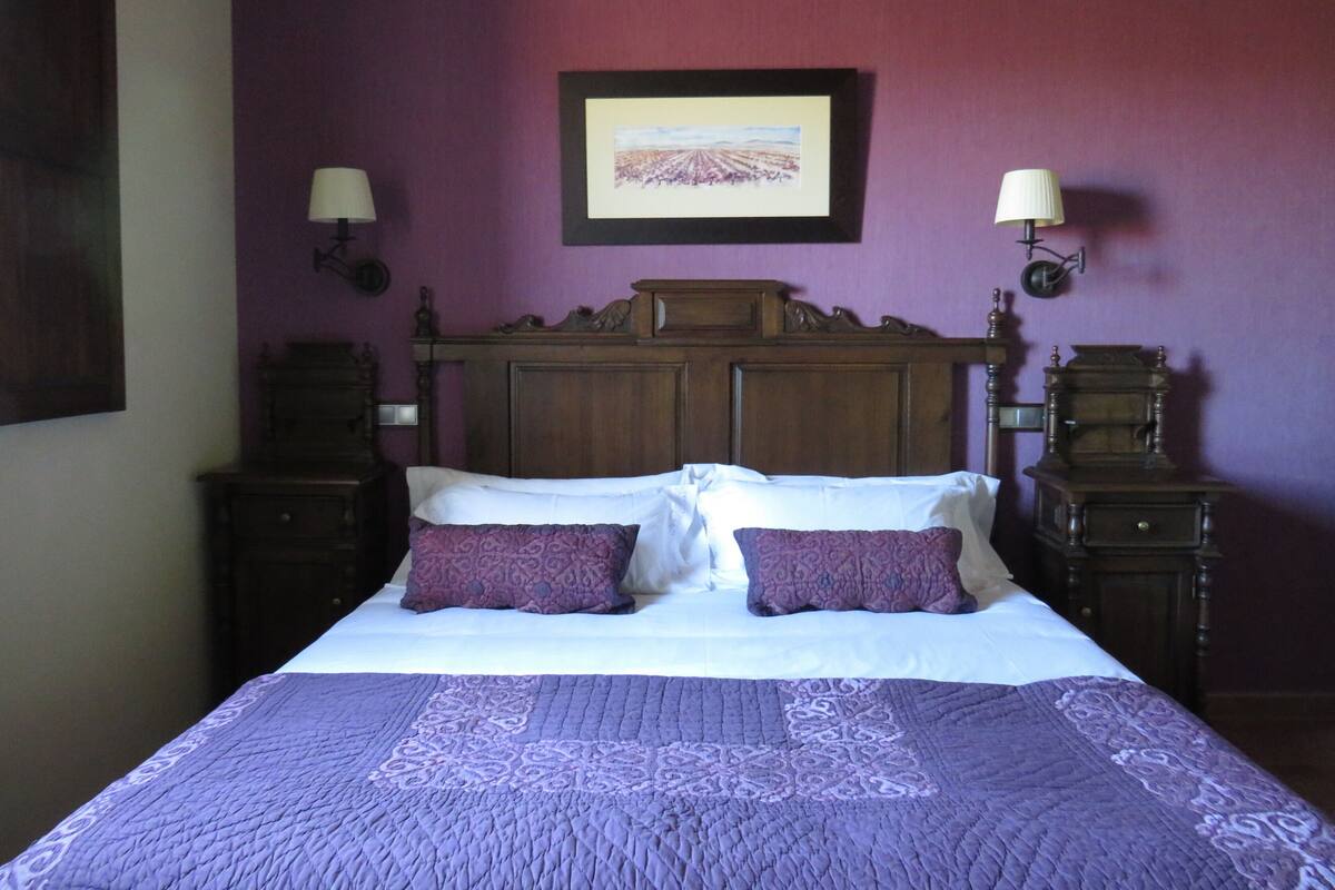 Un dormitorio con cabecera de madera, dos lámparas de noche, una cama con ropa de cama blanca y almohadas moradas, y un cuadro en la pared.
