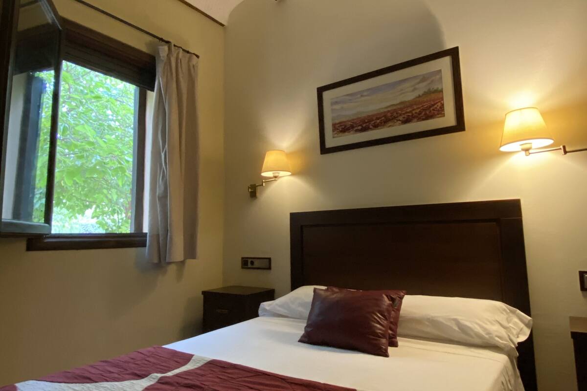 Un dormitorio con una cama, un ventanal, un cuadro y dos lámparas fijadas a la pared.
