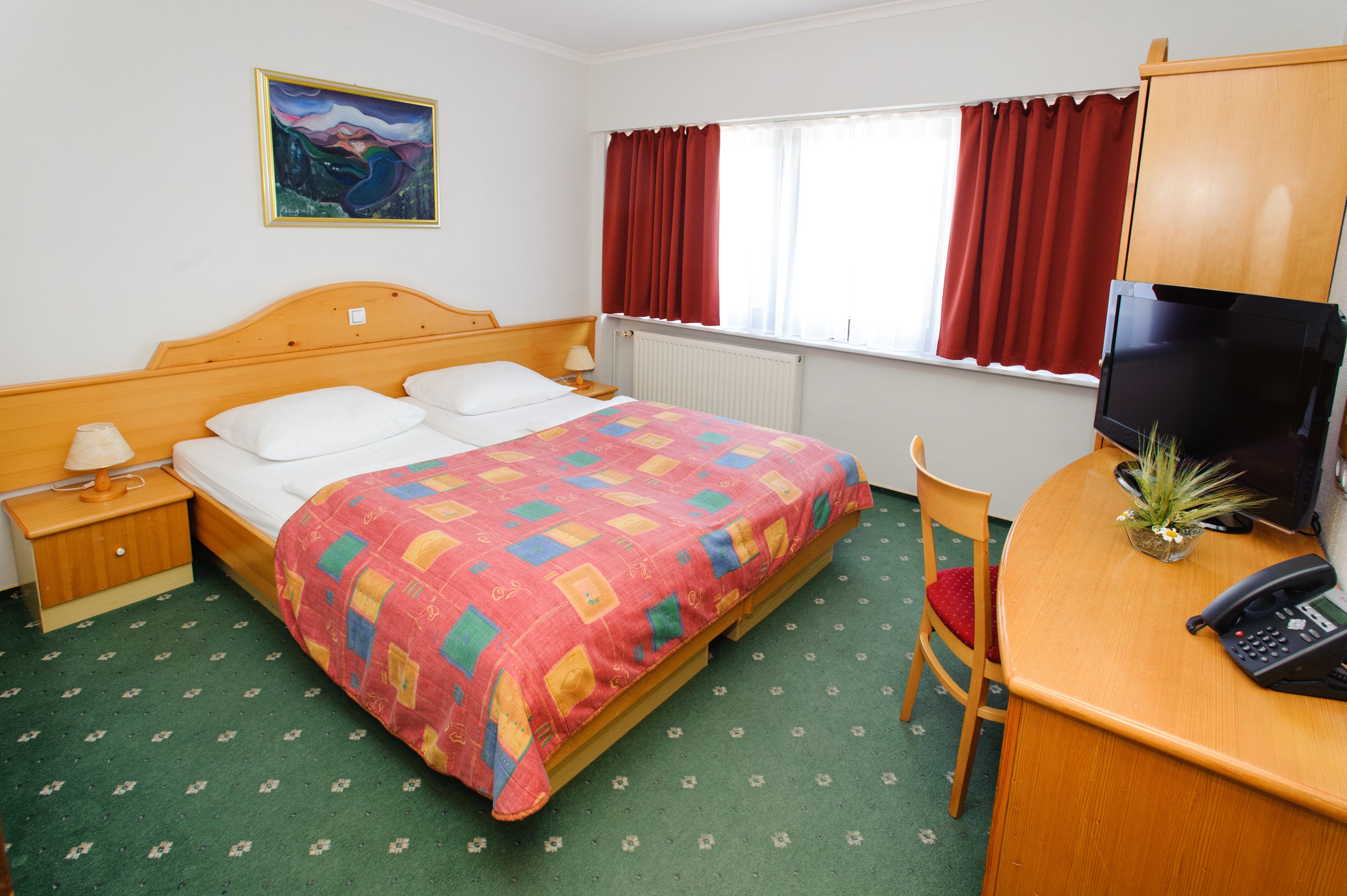 A hotel room with a wooden bed, a desk with a telephone, a chair, a radiator, and a window with curtains.