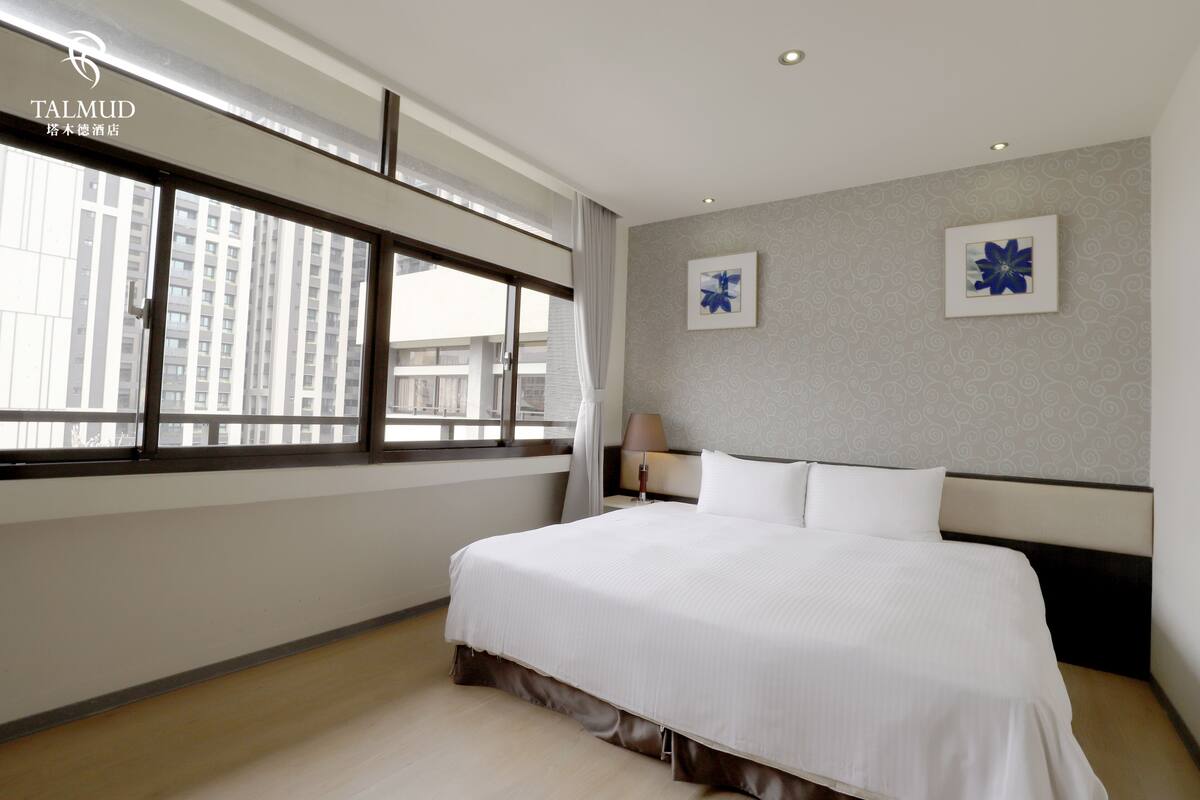 A hotel room with a large bed, a window with curtains, a wall-mounted lamp, and two framed pictures.