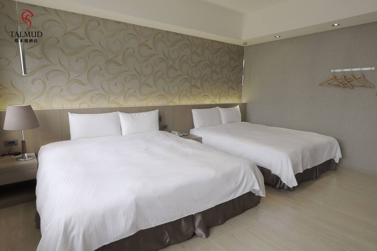 A hotel room with two beds, a nightstand with a lamp, a wall-mounted phone, and a hanging clothes rack.