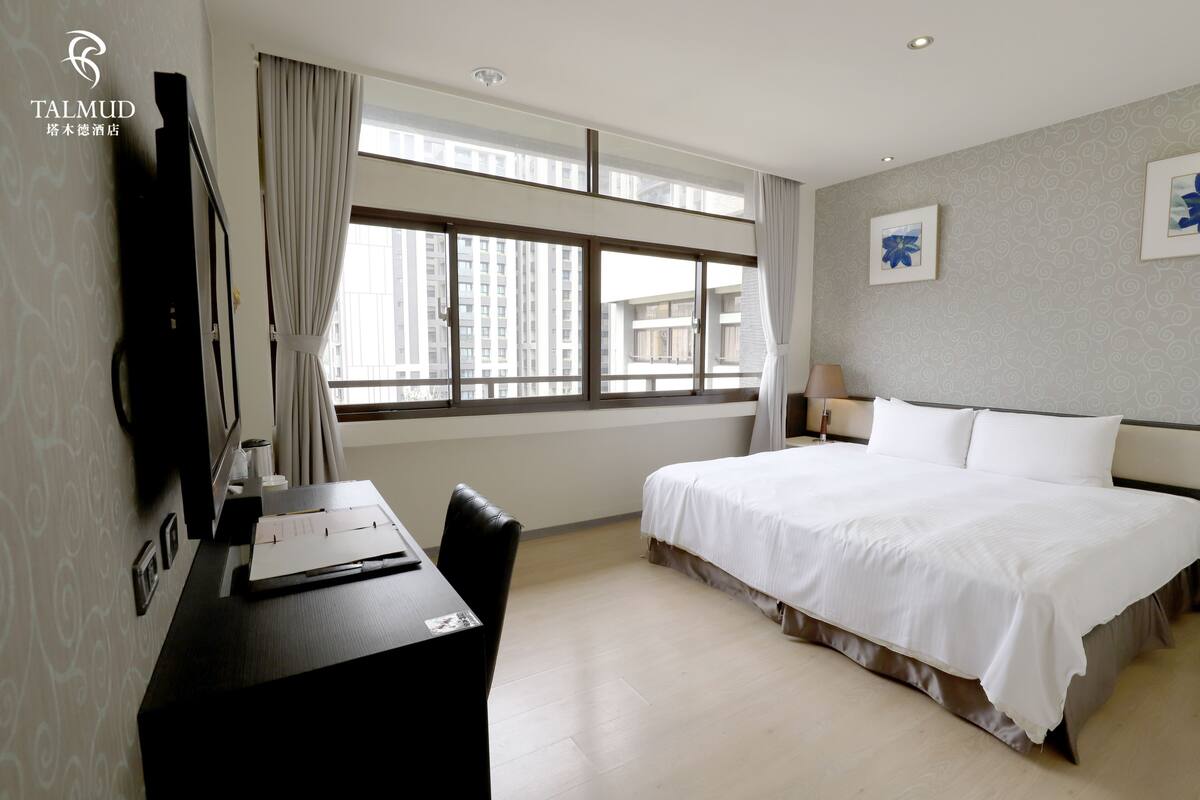 A hotel room with a large bed, a desk with a chair, a TV, and a window with curtains.