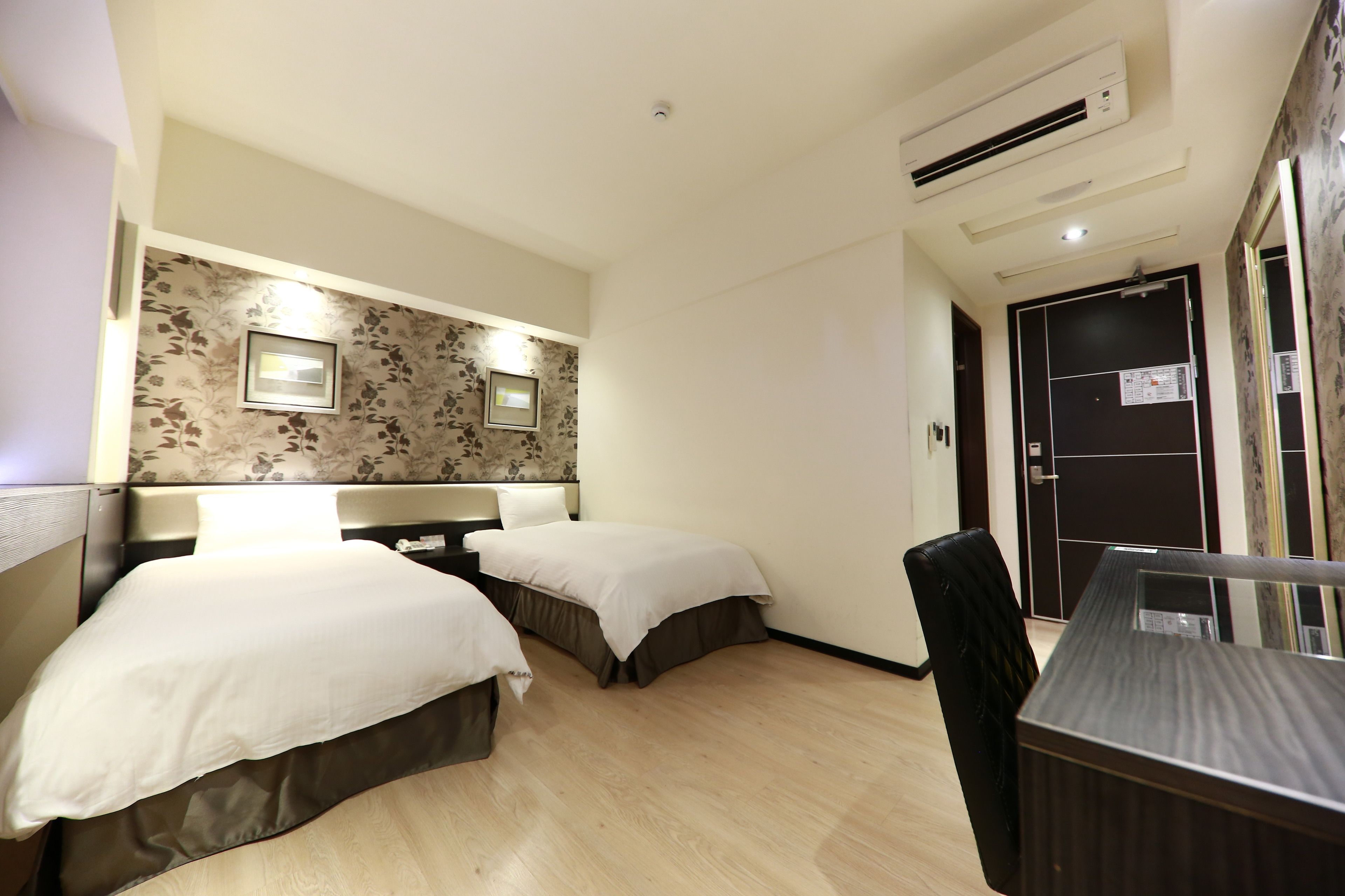 A hotel room with two beds, a desk, a chair, a wardrobe, and an air conditioning unit.