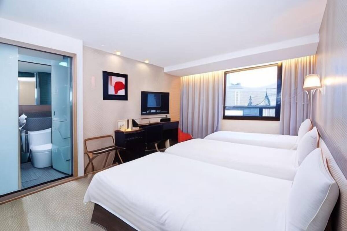 A hotel room with two beds, a desk with a TV, a bathroom, and a window with a city view.