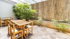 A greenhouse with wooden tables and chairs, surrounded by bamboo plants and a wooden wall.