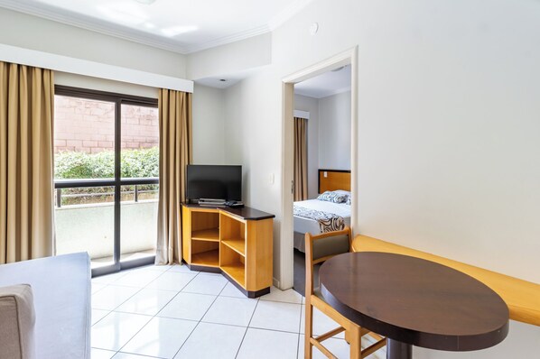 Habitación de hotel con cama, televisor sobre un soporte de madera, una mesita y vistas al exterior.