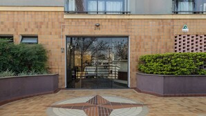 A building entrance with a numbered door, a tiled floor with a geometric pattern, and a small garden area with bushes.