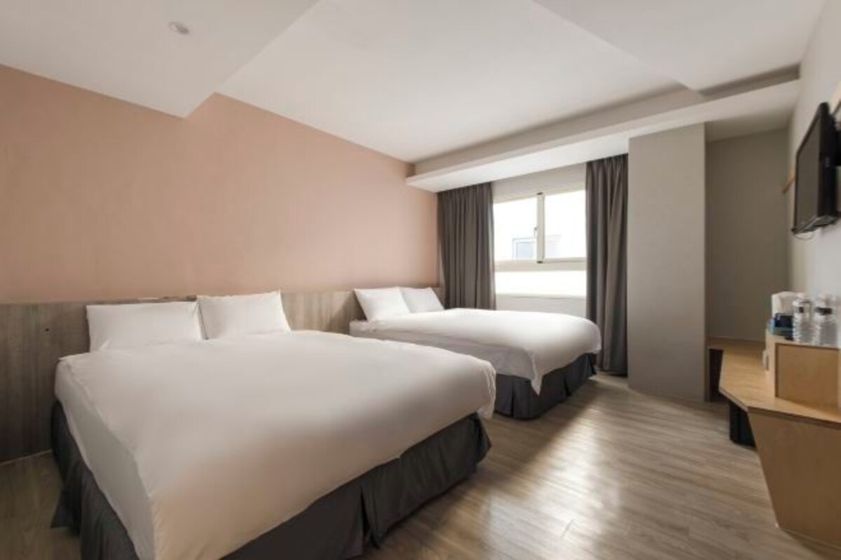 Ein Hotelzimmer mit zwei Betten, einem Holztisch, einem Flachbildfernseher und einem Fenster mit Vorhängen.
