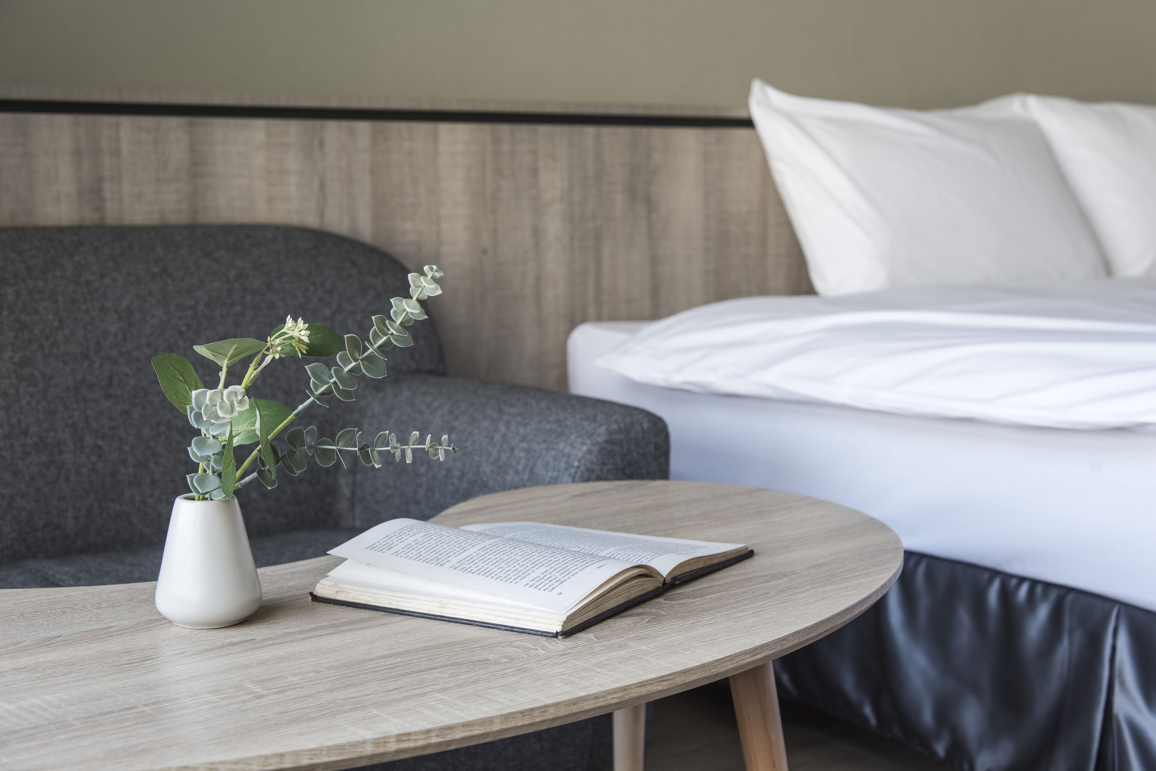 A neatly arranged hotel room with a round wooden table, a white vase with a plant, and a bed with white linens.