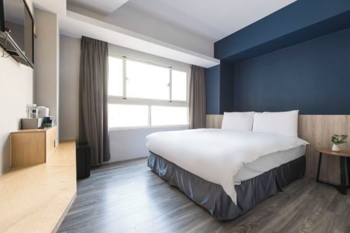 Ein modernes Hotelzimmer mit einem großen Bett, einem kleinen Nachttisch, einem an der Wand montierten Fernseher und einem Fenster mit Vorhängen.