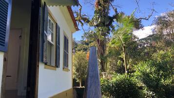 Una casa con fachada blanca, base amarilla y una puerta con ventanal. Se observa un jardín con diversas plantas y un árbol.