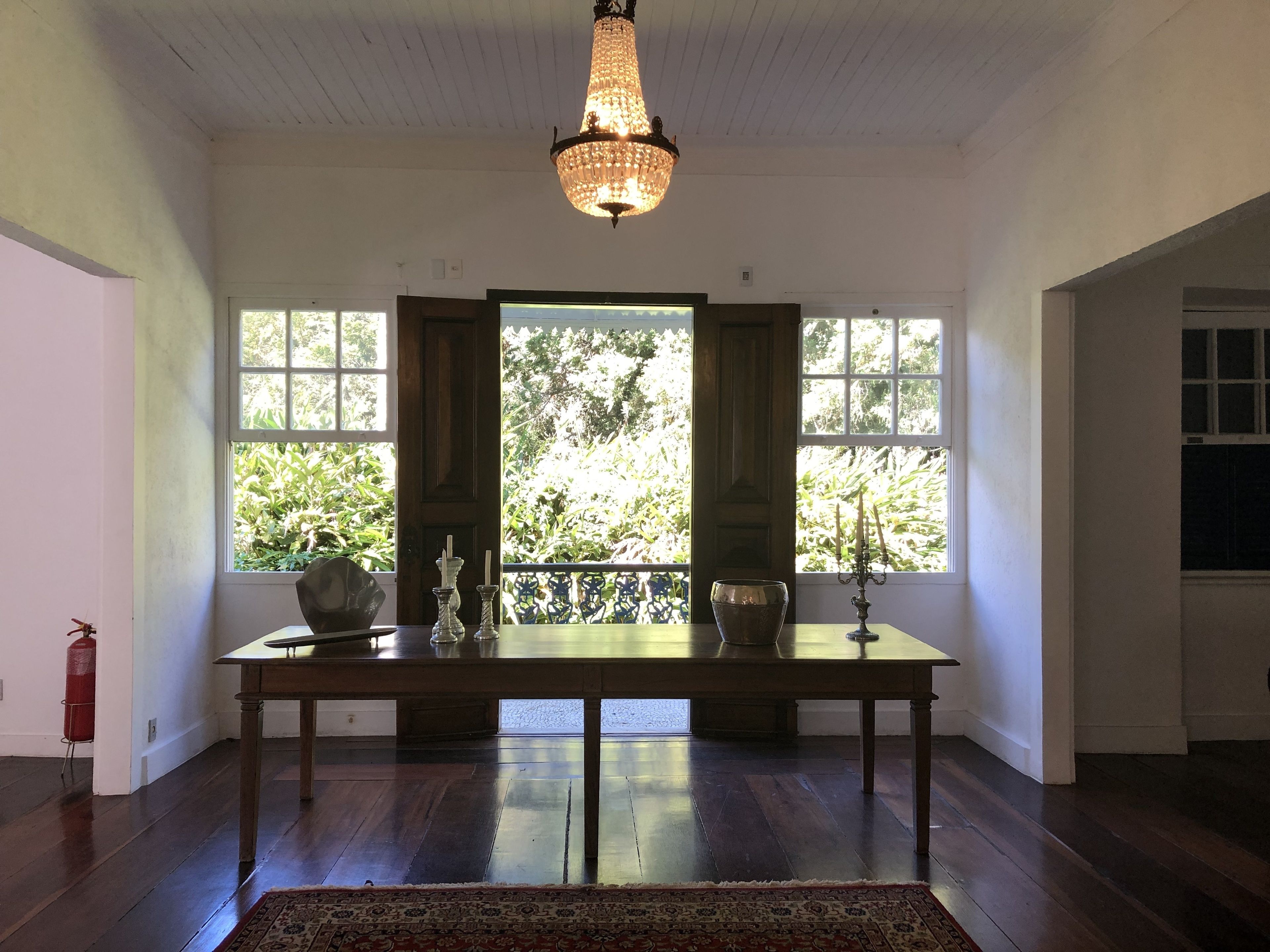 A room with a wooden table, a chandelier, and large windows with a view of greenery.