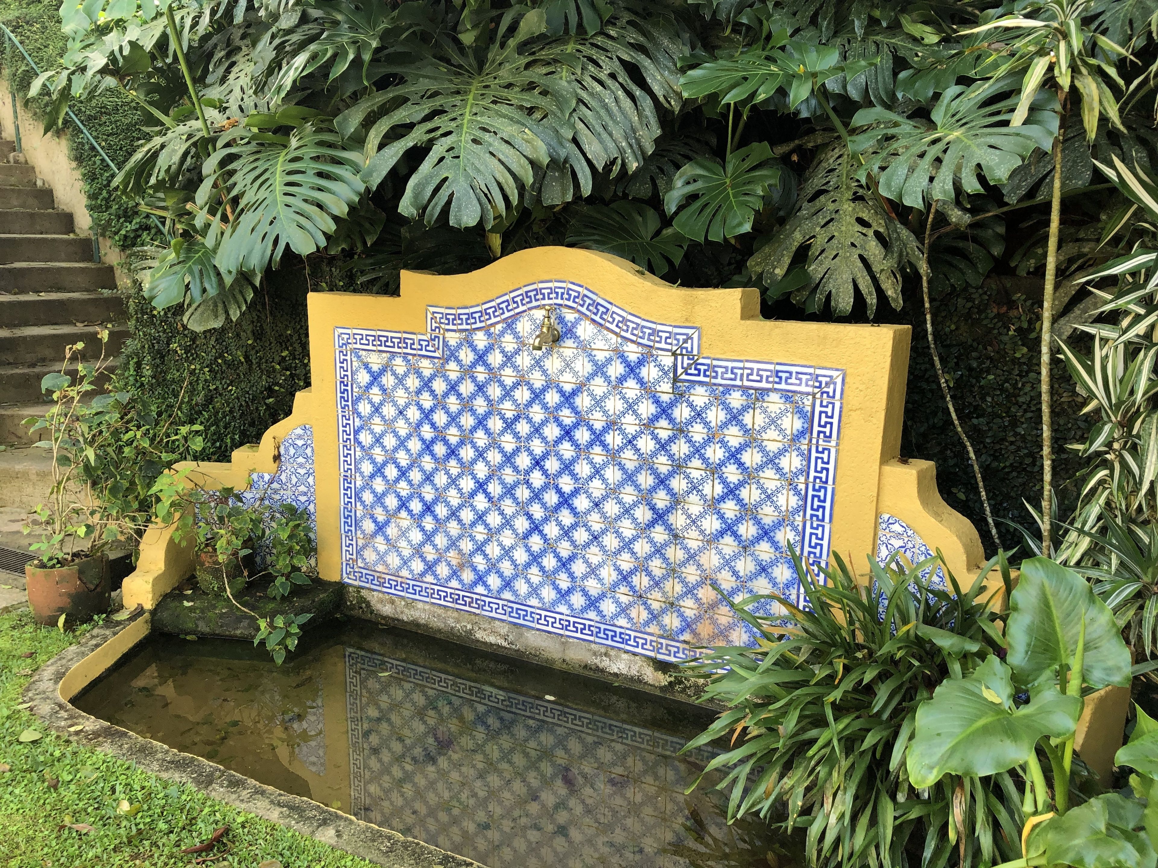 Pared de azulejos con un diseño geométrico, rodeada de abundante follaje y una pequeña fuente de agua.