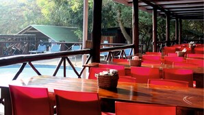 Een overdekt terras met rode stoelen en tafels, met uitzicht op een zwembad en een gebouw met een groen dak.