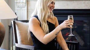 A woman seated in a chair, holding a glass of champagne. She is in a room with a fireplace, a lamp, and a table with a tray of glasses and bottles. - International Hotel Wagga Wagga (East Wagga Wagga)
