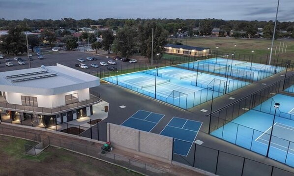 Un complejo deportivo con varias canchas de tenis, una zona de espectadores techada y un estacionamiento.