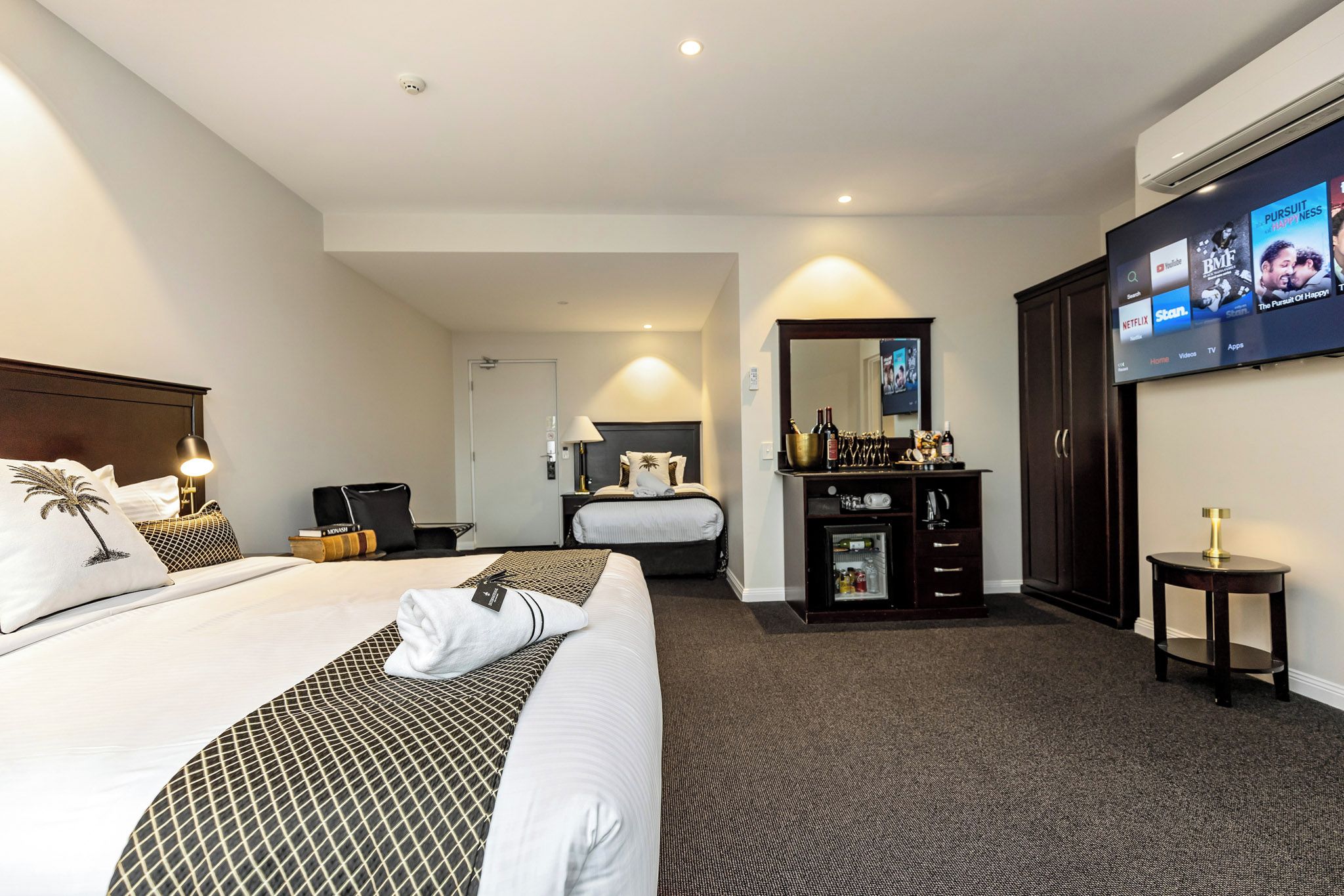 A hotel room with a large bed, a desk with a computer, a minibar, and a television displaying various channels.